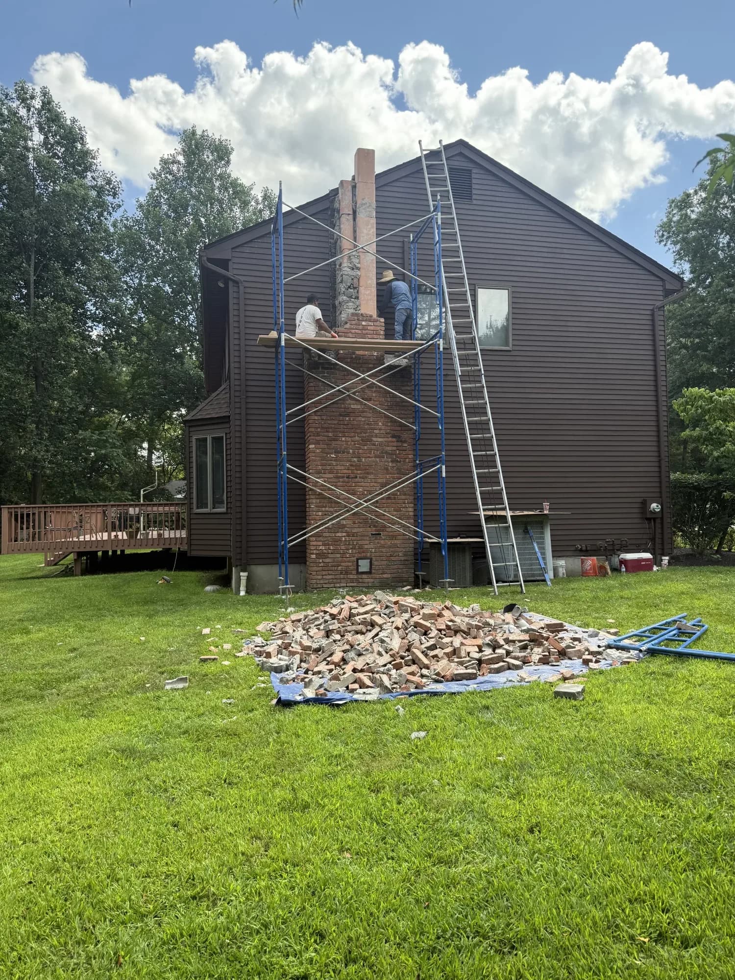 Masonry crew on scaffolding rebuilding a tall brick chimney on a New Jersey home, with the old bricks piled on the lawn below
